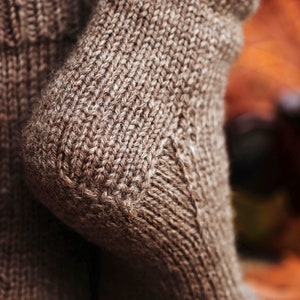 Woman wears The Beginner Socks that were knitted in brown worsted weight yarn using the knitting pattern. This easy knitting pattern lets you knit socks fast and quick, and features a classic knit sock construction and cozy knit stitches all over.