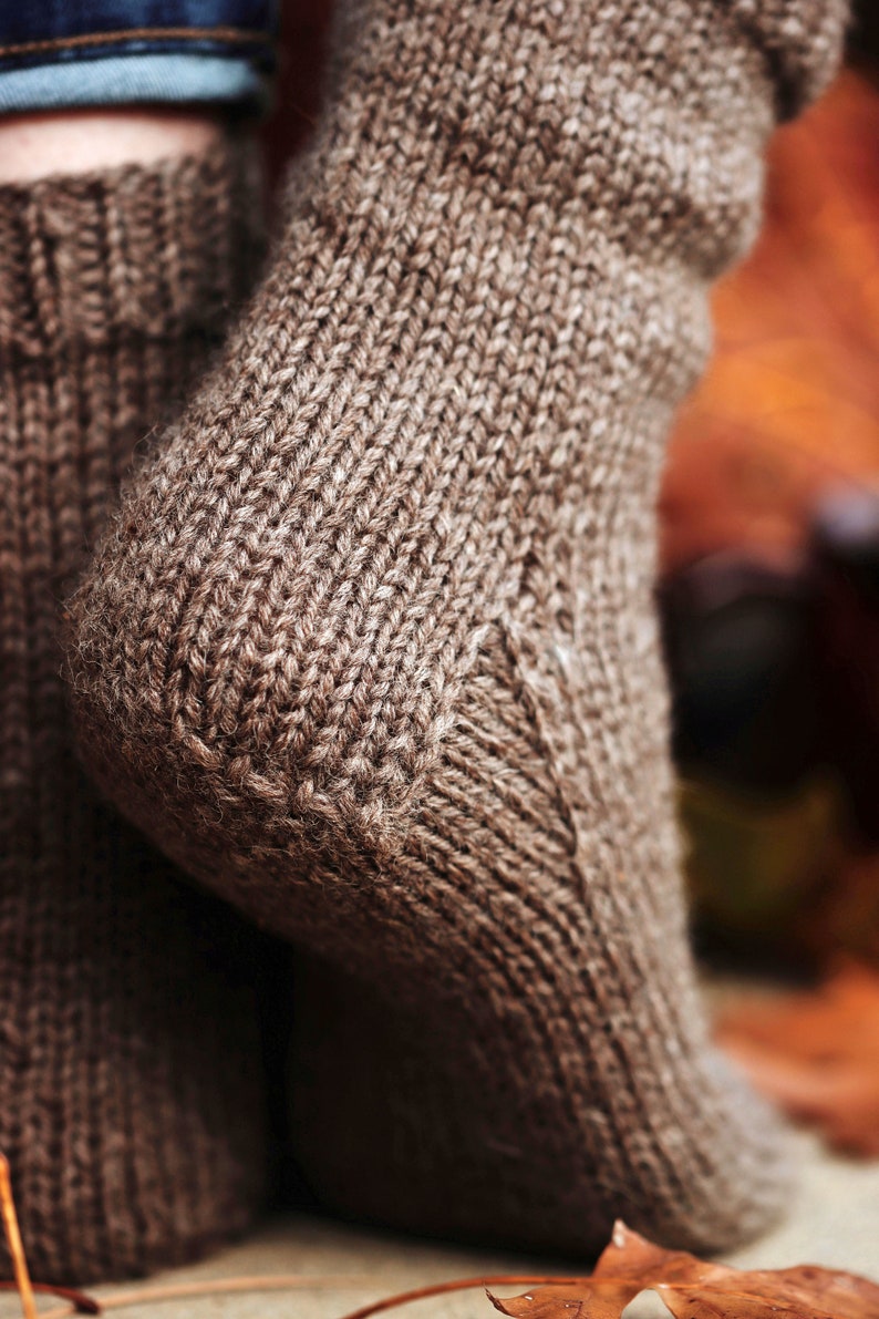 Woman wears The Beginner Socks that were knitted in brown worsted weight yarn using the knitting pattern. This easy knitting pattern lets you knit socks fast and quick, and features a classic knit sock construction and cozy knit stitches all over.