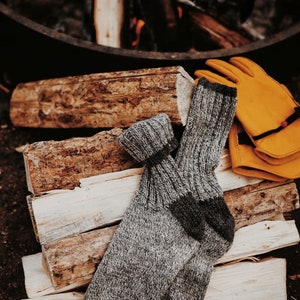 A pair of Camp Socks, a knitting pattern, are laid on a pile of firewood. The socks are knit in two colors of grey wool yarn and these knit boot socks look warm and cozy to wear while camping or hiking. [boot socks knit pattern, socks knit pattern]