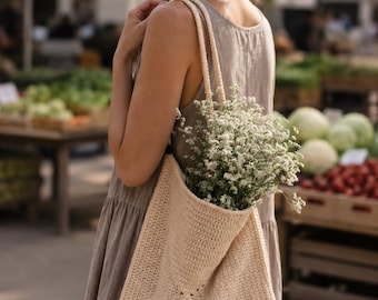 May include: A beige crochet tote bag with two braided shoulder straps, featuring a square-based construction and a textured stitch pattern. The bag is filled with a bouquet of small white flowers, showcasing a simple and functional design.