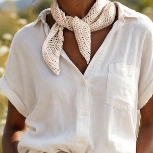 May include: A cream-colored, triangular crochet neck scarf with an open-knit pattern, tied in a knot at the front. The scarf is worn over a short-sleeved, button-down shirt in a light beige tone, featuring a single chest pocket and cuffed sleeves.