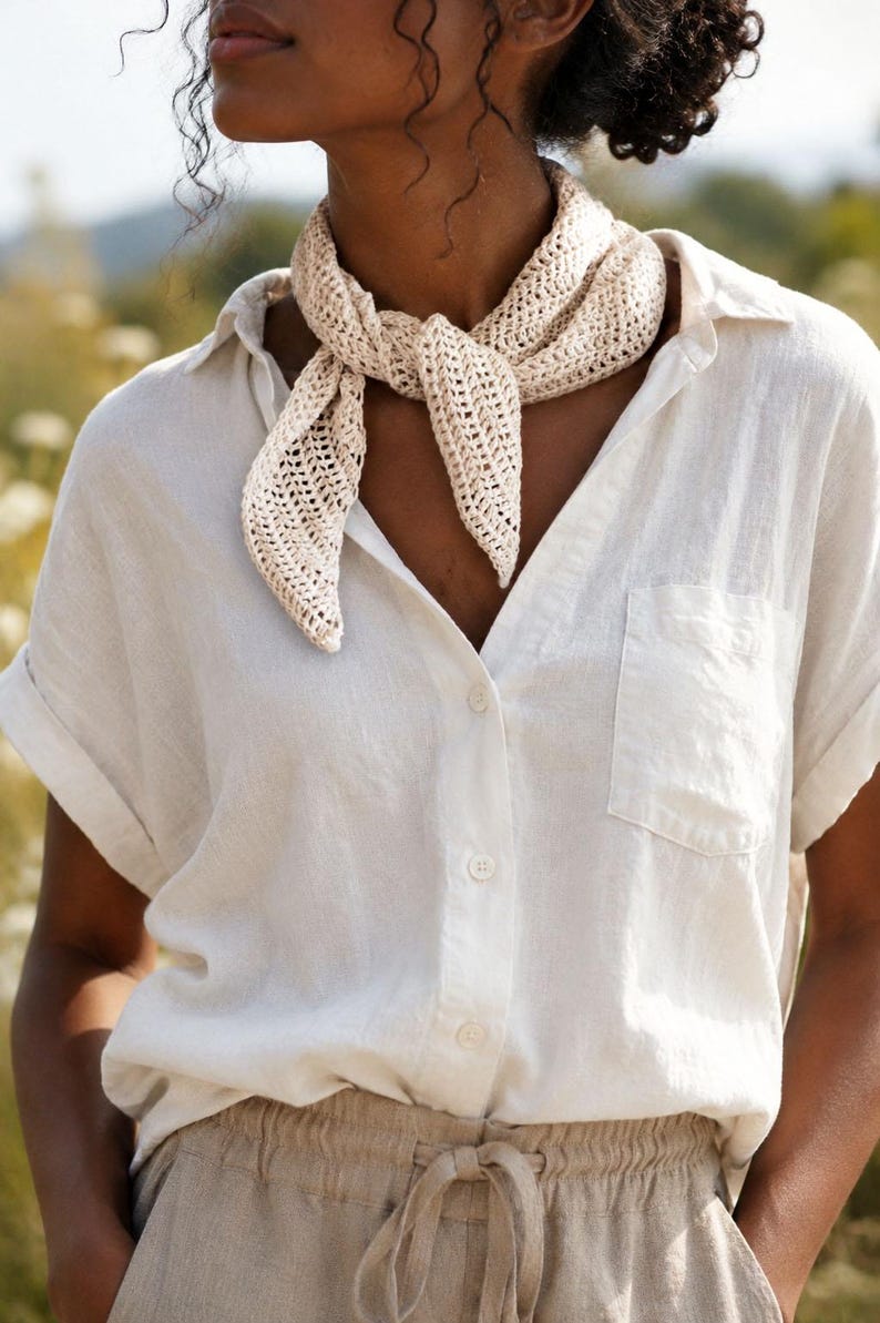 May include: A cream-colored, triangular crochet neck scarf with an open-knit pattern, tied in a knot at the front. The scarf is worn over a short-sleeved, button-down shirt in a light beige tone, featuring a single chest pocket and cuffed sleeves.