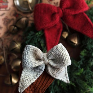 May include: A close-up of a knitted bow in shades of gray and white, with a larger red knitted bow in the background. The bows are set against a backdrop of greenery and holiday decorations, suggesting a festive theme.