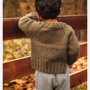Könnte beinhalten: Ein langärmeliger, olivgrüner Strickpullover mit geripptem Saum und Bündchen, kombiniert mit einer hellgrauen Jogginghose. Der Pullover hat einen Rundhalsausschnitt und ein strukturiertes Strickmuster am unteren Rücken, getragen von einem Kind.