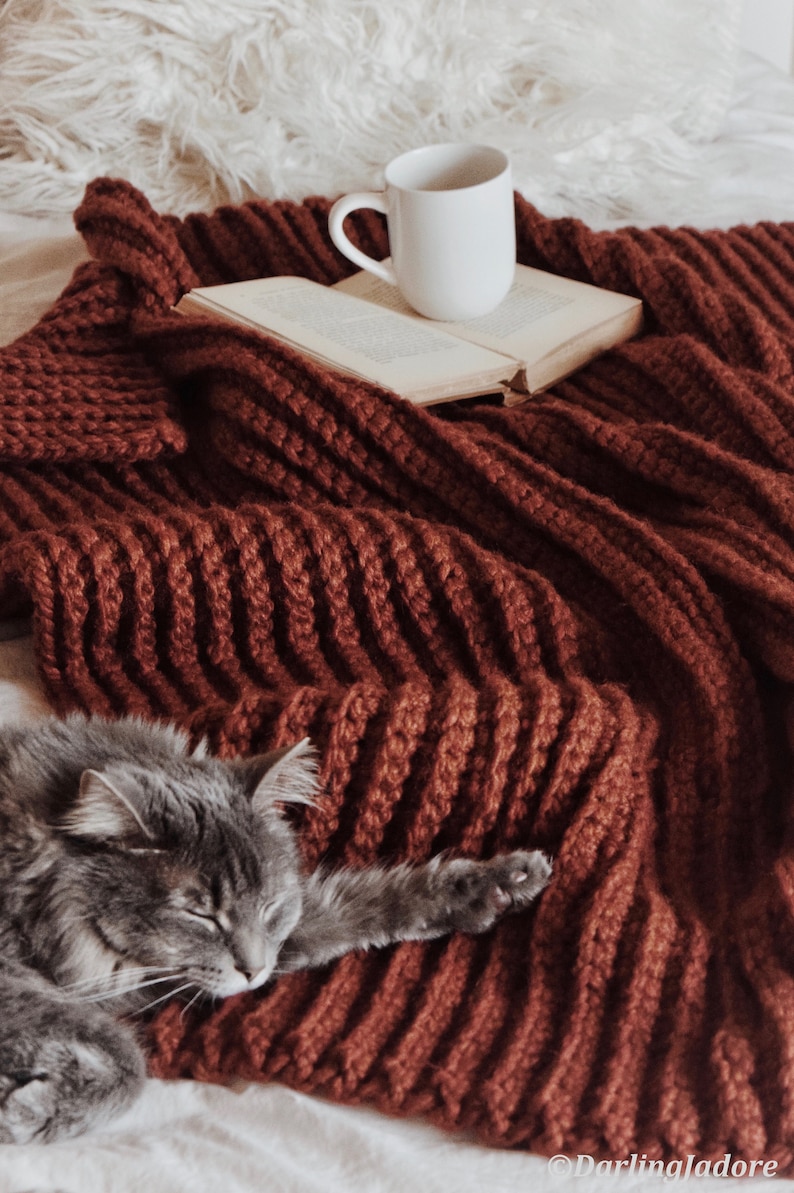 A chunky crochet blanket is shown in a rib fabric with a red yarn. A cat lays on top of the cozy crochet afghan, and it looks very warm and like a pretty home decor item.