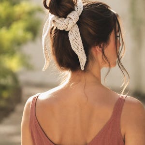 May include: An off-white crochet hair accessory featuring a textured, open-knit pattern. The accessory is tied around a hair bun, with two long, pointed ends hanging down, creating a decorative knot detail that secures the hair in place.