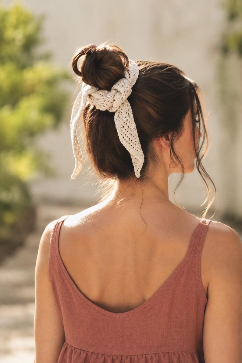 May include: An off-white crochet hair accessory featuring a textured, open-knit pattern. The accessory is tied around a hair bun, with two long, pointed ends hanging down, creating a decorative knot detail that secures the hair in place.