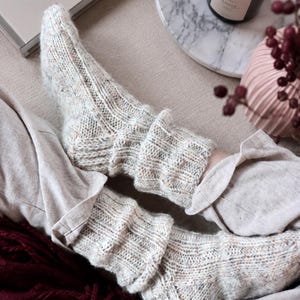 May include: A pair of cream-colored, speckled knit socks featuring a ribbed texture and a folded cuff design. The socks are crafted from a textured yarn with subtle flecks of color throughout, providing a thick, cozy appearance with a visible knit pattern.