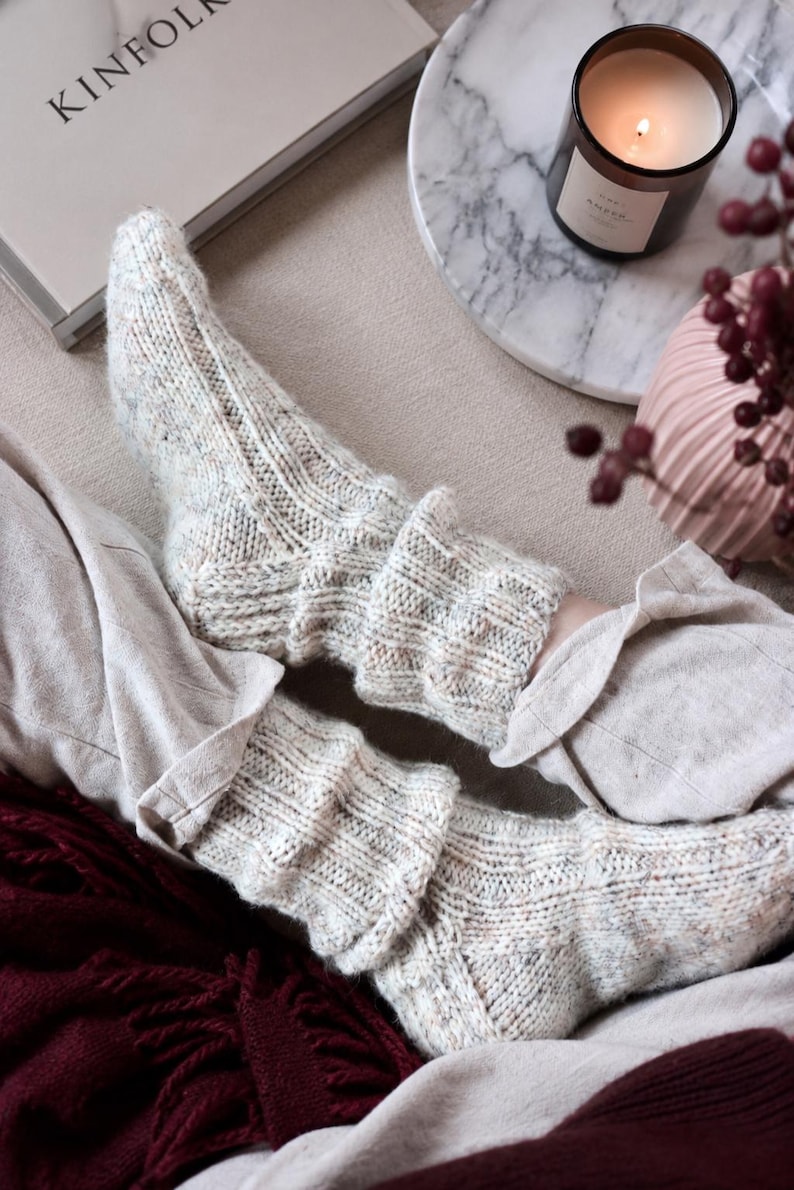 May include: A pair of cream-colored, speckled knit socks featuring a ribbed texture and a folded cuff design. The socks are crafted from a textured yarn with subtle flecks of color throughout, providing a thick, cozy appearance with a visible knit pattern.
