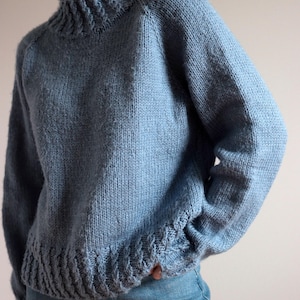 Cable knit sweater pattern, easy raglan sweater knitting pattern. women top down seamless raglan sweater with cables. oversize knit sweater pattern for women. beginner turtleneck pullover sweater knit pattern. high neck jumper knit pattern mock neck.  A light blue knit sweater with a ribbed neckline, cuffs, and hem. The long-sleeved sweater has a relaxed fit and is made of a soft, textured yarn. The sweater is paired with blue jeans.