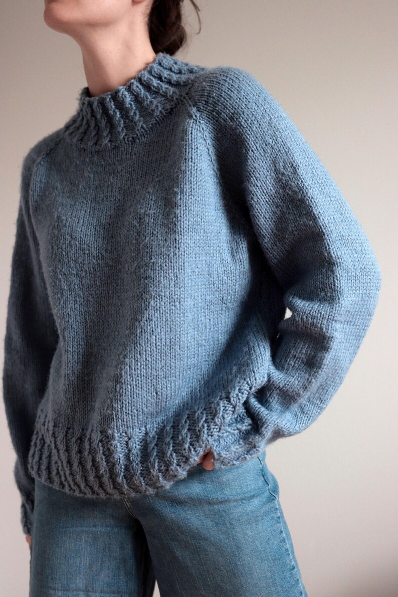 Cable knit sweater pattern, easy raglan sweater knitting pattern. women top down seamless raglan sweater with cables. oversize knit sweater pattern for women. beginner turtleneck pullover sweater knit pattern. high neck jumper knit pattern mock neck.  A light blue knit sweater with a ribbed neckline, cuffs, and hem. The long-sleeved sweater has a relaxed fit and is made of a soft, textured yarn. The sweater is paired with blue jeans.