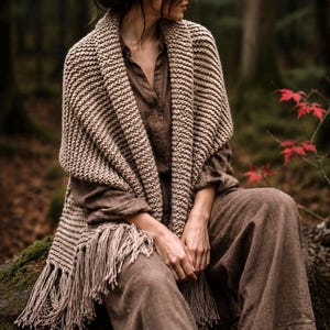 May include: A beige knitted shawl featuring a thick garter stitch texture and long fringe trim along the bottom edges. The shawl is draped over a brown long-sleeved button-down shirt and matching wide-leg trousers made of a textured fabric.