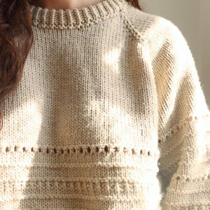May include: A beige long-sleeve sweater featuring a ribbed crew neckline and raglan sleeves. The knit garment displays a textured pattern with horizontal rows of eyelet cutouts and raised stitch details across the chest and sleeves.