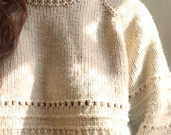 May include: A beige long-sleeve sweater featuring a ribbed crew neckline and raglan sleeves. The knit garment displays a textured pattern with horizontal rows of eyelet cutouts and raised stitch details across the chest and sleeves.