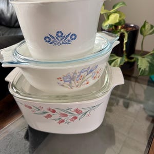 May include: Three white CorningWare casserole dishes with glass lids are stacked. The dishes have floral patterns in blue, pink, and purple. The dishes are on a glass table.