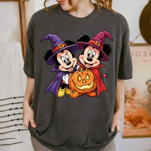 May include: A charcoal gray t-shirt featuring a graphic of two cartoon mice dressed as witches with purple and red capes and pointed hats. They are standing behind a large orange jack-o'-lantern pumpkin. A small black spider is visible to the right.