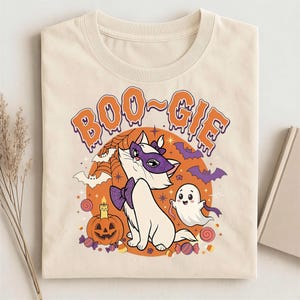Op de afbeelding: Een crèmekleurig t-shirt met een afbeelding van een witte kat met een paars masker en strikje, een klein spookje, een pompoen, vleermuizen en spinnenwebben. De tekst "BOO-GIE" is in oranje met een druipend effect boven de illustratie gedrukt.