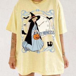 May include: A pale yellow t-shirt featuring a graphic of a blonde character in a light blue gown, black cape, and witch hat. The character holds an orange jack-o'-lantern. The design includes a clock face, bats, a skull, and the text SPOOKY PRINCESS.