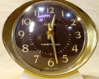 Puede incluir: Un despertador Westclox con esfera redonda de color marrón oscuro, números dorados y manecillas doradas. El reloj está encerrado en un marco de metal dorado pulido y se asienta sobre una base rectangular blanca. El texto "Westclox" y "Made in U.S.A." aparece en la esfera.
