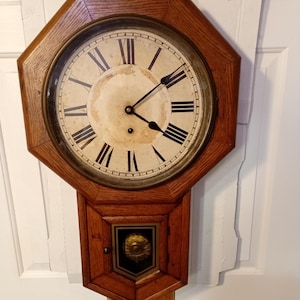 May include: Antique wooden wall clock with an octagonal frame. The clock face features black Roman numerals and hands, with a faded cream-colored background. The lower section has a decorative panel and pendulum rods.