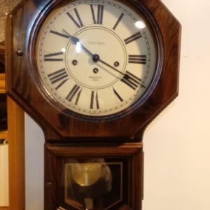 May include: A vintage wall clock with a dark wood octagonal frame. The clock face is cream-colored with black Roman numerals and hands. The clock has a pendulum visible through a glass window.