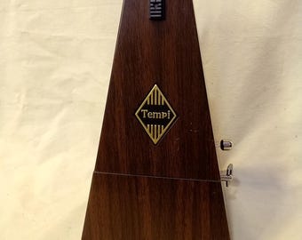 Puede incluir: Un metrónomo de madera de color marrón oscuro con veteado y forma piramidal. Presenta un deslizador rectangular negro cerca de la parte superior y un emblema dorado en forma de diamante en el centro con la palabra Tempi impresa en negro.