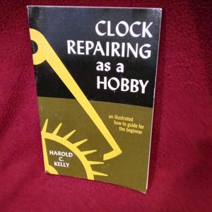 May include: A paperback book titled Clock Repairing as a Hobby by Harold C. Kelly. The cover features a black top section with white text and a dark olive green bottom section with a yellow graphic of a clock gear and hand, plus subtitle text.