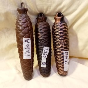 May include: Three antique, brown, pinecone-shaped clock weights. Each weight has a loop at the top and a label with the number "1260" and a letter. The weights are displayed on a cream-colored surface.