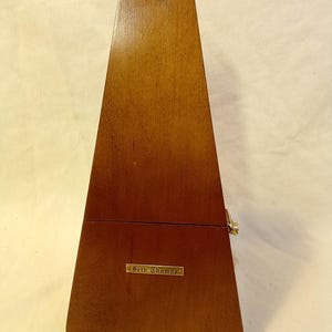 May include: A wooden metronome with a pyramid shape. The metronome is a warm brown color and has a small brass plate that reads "Seth Thomas". The top of the metronome has a small, triangular shape.