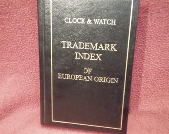 Puede incluir: Un libro de tapa dura negro con letras doradas en la portada que dice CLOCK & WATCH TRADEMARK INDEX OF EUROPEAN ORIGIN. La cubierta presenta un borde rectangular dorado fino que enmarca el texto, con un acabado texturizado en la superficie.
