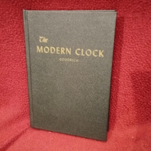 May include: A black hardcover book titled The Modern Clock by Goodrich. The front cover features a textured finish with the title and author name printed in gold lettering. The book is positioned vertically, showcasing the front cover design.