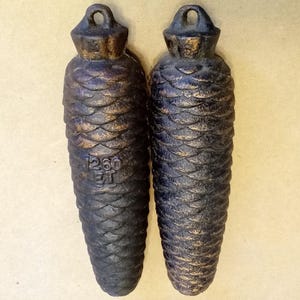 May include: Two dark, heavy cast iron clock weights shaped like pine cones. Each weight features a textured, overlapping scale pattern and a circular hanging loop at the top. The left weight is embossed with the text 1260 ET, while the right weight has a small number 5 visible near the top loop.