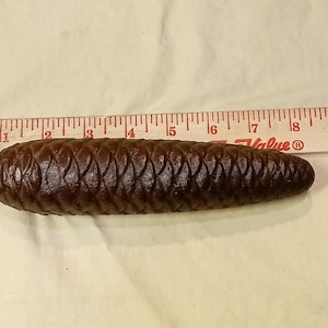May include: A brown, pinecone-shaped clock weight with a metal loop for hanging. The weight is approximately 6 inches long and is next to a wooden ruler.