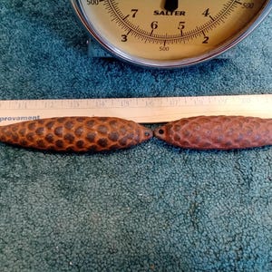 May include: Two brown, textured, cast iron, pine cone shaped doorstops, each measuring approximately 14 inches in length, are placed side-by-side on a blue, fuzzy surface. A wooden ruler with the words "Home Improvement" printed on it is placed behind the doorstops.