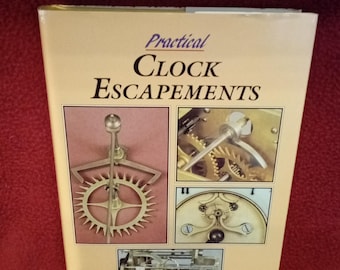Puede incluir: Un libro de tapa dura titulado Practical Clock Escapements de Laurie Penman. La cubierta de color crema presenta tres recuadros fotográficos distintos que muestran varios mecanismos de escape de reloj en latón, incluyendo engranajes y palancas.