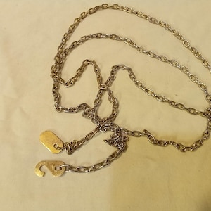 May include: A vintage gold-toned chain belt with a hook closure and a small rectangular tag. The chain is composed of oval links and is displayed on a light-colored surface. The belt is likely from the mid-century.