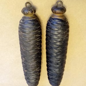 May include: Two dark, weathered cast iron weights shaped like pine cones. Each weight features a textured, overlapping scale pattern and a rounded top with a small metal loop for hanging. The finish is a deep, matte black with subtle brown tones.