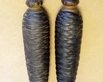 Puede incluir: Dos pesas de hierro fundido oscuras y desgastadas con forma de piña. Cada pesa presenta una textura de escamas superpuestas y una parte superior redondeada con una pequeña anilla metálica para colgar. El acabado es negro mate con tonos marrones.