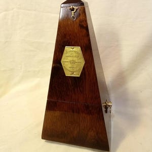 May include: A dark brown wooden metronome with a pyramid shape. It features a brass hexagonal plaque on the front engraved with the text METRONOME DE MAELZEL, a small brass hook at the top, and a brass latch on the side for securing the casing.