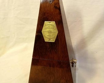 Puede incluir: Un metrónomo de madera marrón oscuro con forma de pirámide. Presenta una placa hexagonal de latón en la parte frontal grabada con el texto METRONOME DE MAELZEL, un pequeño gancho de latón en la parte superior y un cierre de latón en el lateral.