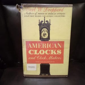 May include: A yellow book cover with the title "American Clocks and Clock Makers" in red and black lettering. The book cover features a black and white illustration of an antique clock with a Roman numeral clock face. The book is by Carl W. Drepperd, author of "Primer of American Antiques" and "First Reader for Antique Collectors".