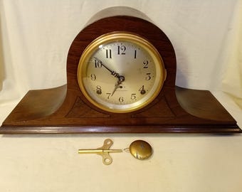 Puede incluir: Un reloj de chimenea de madera con esfera y agujas doradas, con números romanos. El reloj tiene una parte superior curvada y una base rectangular. Una llave y un péndulo dorados están colocados delante del reloj.