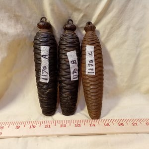 May include: Three antique clock weights with a pinecone design. The weights are dark brown, black, and brown, with white labels marked "1170 A", "1170 B", and "1170 C". A wooden ruler is at the bottom.