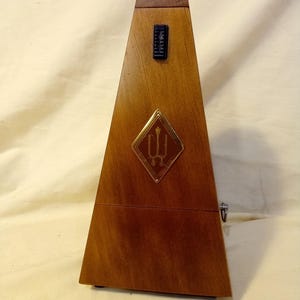 May include: A wooden metronome in a pyramid shape. The metronome is brown with a gold-colored emblem and a black scale. The metronome has a hinged top and a pendulum. A classic tool for musicians.