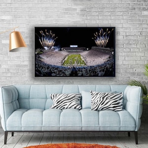 May include: A framed stadium print featuring a nighttime football game with fireworks. The stadium is filled with spectators, and the field is illuminated. The print hangs above a light blue couch with zebra-print pillows.