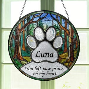 Puede incluir: Vidriera redonda con una escena de bosque y un diseño de huella de pata. La huella de pata tiene el nombre "Luna" y el texto "You left paw prints on my heart". La obra de arte está enmarcada con un borde de metal negro y cuelga de una cadena.
