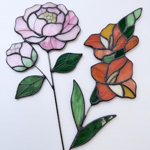 May include: Two stained glass flower sculptures. One features a pink peony with a bud and green leaves. The other is an orange and yellow lily with green leaves. Both have black metal stems.