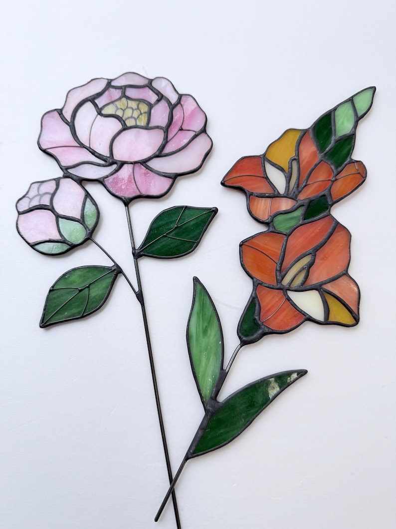 May include: Two stained glass flower sculptures. One features a pink peony with a bud and green leaves. The other is an orange and yellow lily with green leaves. Both have black metal stems.