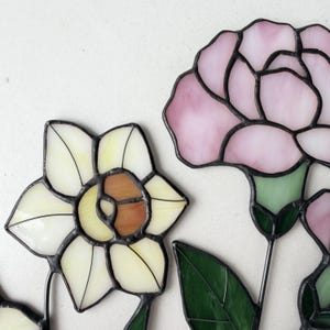 May include: Close-up of stained glass flowers, including a daffodil with white and brown petals and a pink carnation. The flowers have black outlines and green leaves. The glass is translucent.