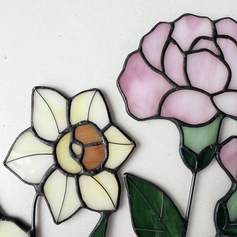May include: Close-up of stained glass flowers, including a daffodil with white and brown petals and a pink carnation. The flowers have black outlines and green leaves. The glass is translucent.
